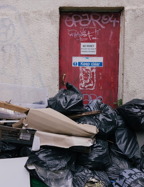 A collection of black garbage bags filled with waste piled against a weathered red door set into a plain, textured beige wall. The door features various graffiti tags in white and pink spray paint, along with stickers including one that says 'Keep clear' and another indicating 'NO MONEY' with additional smaller text. Surrounding the bags are loose household items such as flattened cardboard boxes, a piece of foam or padding, and a small metal or plastic container partially visible on the left side. The scene is outdoors, likely on a paved or concrete surface, and appears to be a designated area for rubbish accumulation, possibly related to non-sanctioned or independent waste disposal, as suggested by the clutter and the context of rubbish removal services like those provided by House Clearance Harrow. Natural lighting highlights the textures of the bags, the rough surface of the wall, and the graffiti, conveying a scene that underscores the need for proper waste management or private rubbish collection rather than standard municipal clearance.
