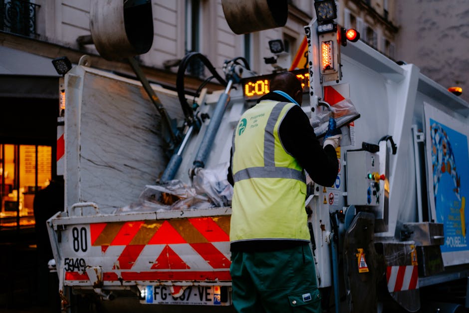 A waste collection worker wearing a high-visibility yellow vest and dark clothing is standing beside a large white rubbish truck with red and orange reflective stripes. The worker is operating the vehicle's control panel, possibly managing the sorting or disposal process. The back of the truck is open, revealing a collection of mixed waste materials, including paper, plastic, and other debris, piled inside. The scene is set outdoors, possibly in an urban environment, with a building featuring lit windows visible in the background. The lighting suggests early evening or dusk. The truck has various safety and operational lights, and the license plate is partially visible. This scene exemplifies on-site waste collection or alternative rubbish handling methods that may be complemented by private rubbish removal services such as those offered by House Clearance Harrow, facilitating efficient rubbish removal without relying solely on local authority collection.