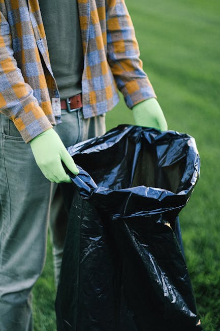 A person holding open a large black plastic rubbish bag outdoors on a grassy area. The individual is wearing a green long-sleeved shirt and a plaid jacket with shades of yellow, gray, and brown. They are also wearing light-colored pants secured with a brown belt and green gloves, indicating they are engaged in waste collection or disposal. The open bag appears empty, ready to be filled with rubbish, and the scene suggests a cleanup activity that could relate to private waste handling or outdoor rubbish removal services. The grassy background and natural lighting highlight the outdoor setting, emphasizing the context of rubbish collection away from public waste authorities, aligning with the concept of independent or off-site waste clearance handled by companies like House Clearance Harrow.