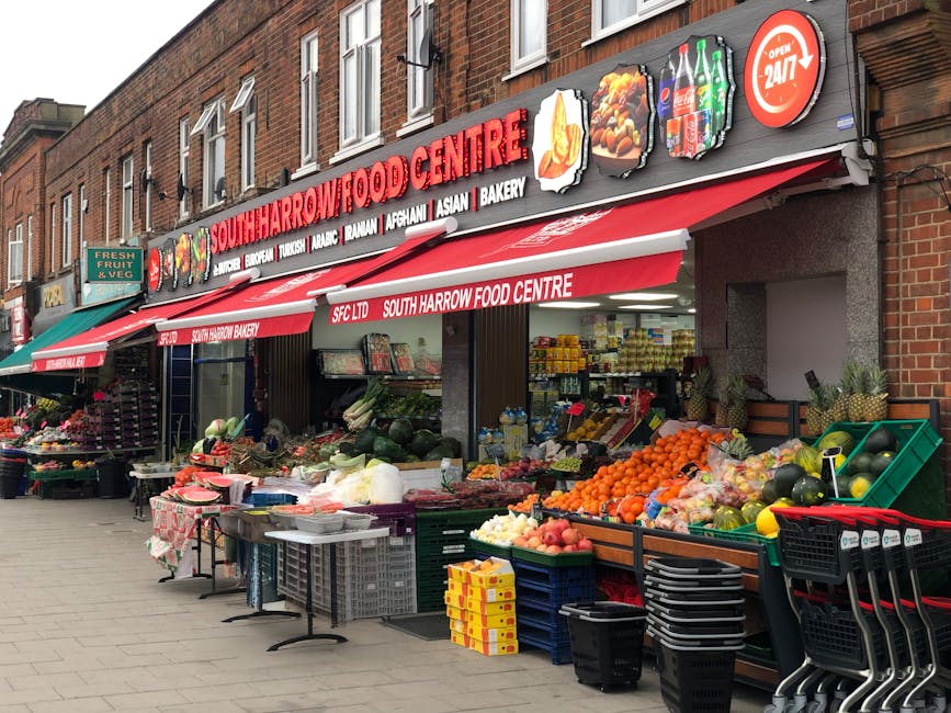 The image depicts the storefront of South Harrow Food Centre, a corner shop situated on a brick building with a multi-pane window on the upper floor. The shop features a red and white signage displaying the shop's name in large, bold letters, along with icons of food items such as a chicken leg, a bottle, and baked goods, indicating its offerings. A red retractable awning extends over the sidewalk, providing shade to the produce displays beneath. On the sidewalk outside, there are several tables and crates filled with an assortment of fresh fruits and vegetables, including oranges, melons, apples, and peppers, arranged in an organized manner. The produce is housed in plastic baskets, crates, and on display tables, some covered with patterned cloths. To the left of the shop, a small green sign indicates the sale of fresh fruit and vegetables, and another awning extends over neighboring shops. The scene is illuminated by daylight, with the storefront appearing clean and well-maintained, capturing the typical setup of a local market or shop that handles private food sales and local produce, fitting into the context of alternative waste management and relevant rubbish removal services such as those offered by House Clearance Harrow.