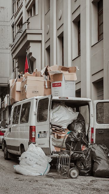 A white panel van parked on a city street with its rear open, revealing an overflowing cargo area filled with large black garbage bags, cardboard boxes, and loose waste materials. Several of the cardboard boxes are stacked on top of the van, some partially crushed or torn, with printed labels and plain brown surfaces. The black rubbish bags appear to be tightly tied, with some resting against the open rear door and others on the ground nearby. A small metal trolley and a wheeled cart are positioned next to the van, loaded with additional black rubbish bags and debris. Behind the vehicle, a multi-storey building with a light grey stone façade features evenly spaced rectangular windows, some with metal grilles, and architectural detailing such as protruding ledges and decorative elements. The scene captures a typical urban environment where private waste collection or rubbish removal services, like those provided by House Clearance Harrow, might be involved in on-site clearance or alternative disposal arrangements, emphasizing the messy accumulation of waste awaiting collection.