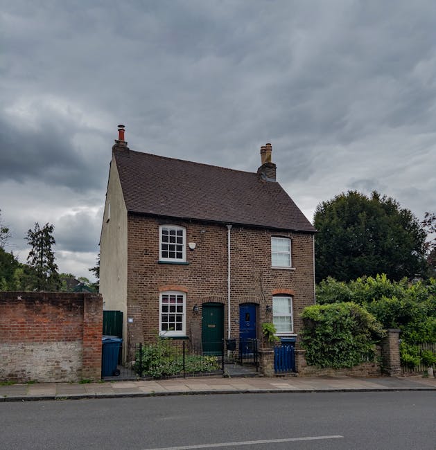 A two-storey brick house with a pitched dark tiled roof and two chimneys, situated on a residential street. The front façade features two windows on the upper floor, each with white frames and sash panes, and two additional windows on the ground floor. The left window has a brick arch detail, while the right one is partially obscured by a green shrub. The front door is located centrally, painted dark blue, and flanked by small garden areas with low metal railings, containing various plants and shrubs. To the left of the house, there is a brick wall with a green gate, and blue wheelie bins are positioned beside the wall. The street in front has a paved sidewalk and a visible road surface, with grey overcast skies overhead. The surrounding environment includes mature trees and bushes, contributing to the suburban setting, which suggests the property may be part of a private residence where private waste collection or on-site clearance could be relevant, aligning with the context of alternative waste handling methods as briefly referenced in the associated service content.