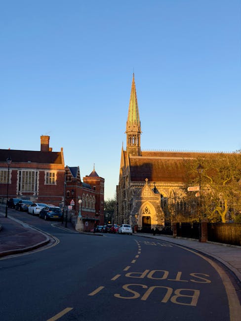 A narrow street in a historic town features a large Gothic-style church with a tall, pointed spire made of weathered stone, situated on the right side of the image. The church's intricate architectural details, including pointed arches and decorative stonework, are visible beneath the sunlight. To the left, red-brick buildings with pitched roofs and chimney stacks line the street, with several parked cars along the curb. The road curves gently to the left, marked by a yellow dashed line and a prominent 'BUS STOP' sign painted on the asphalt. The sky above is clear and blue, casting warm, natural light that highlights the textures of the stone and brick surfaces and produces soft shadows on the buildings and pavement. The environment suggests a quiet, historic area where alternative waste handling, such as private rubbish collection, may be required for local property cleanups, aligning with the context of rubbish removal services like those offered by House Clearance Harrow in managing local waste.