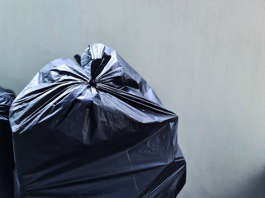 A close-up view of several black rubbish bags made from glossy, plastic material, tightly sealed and stacked against a plain, neutral-colored indoor wall. The bags, which appear to contain household waste or debris, are placed on a flat surface, possibly a floor or a pavement area near a house or property entrance. The bags show creases and subtle reflections of light on their shiny surface, indicating they are filled with soft refuse. Surrounding environment is minimal and uncluttered, with no additional objects or background details visible. This scene reflects typical waste disposal practices where private waste collection or on-site clearance might be used, aligning with independent rubbish removal services such as those offered by House Clearance Harrow. The overall composition emphasizes the readiness of waste for disposal, fitting into context of rubbish removal or bulky waste management in residential settings.