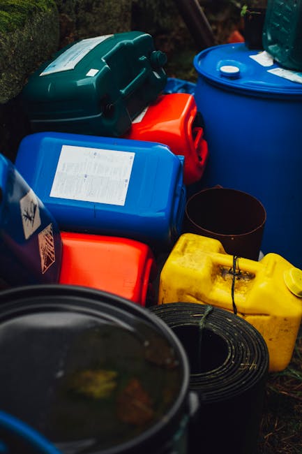 The image shows a collection of various plastic containers and drums placed outdoors on a paved or gravel surface, with some greenery in the background. The containers include a large blue drum with a white lid, a smaller blue container with a label affixed to the top, a bright yellow jerry can, and a green plastic toolbox or storage box nearby. A black coiled hose or cable is also visible in the foreground. The containers appear to be used for storing liquids, likely related to household or garden waste management, and are arranged in a somewhat cluttered manner. This scene reflects a typical setup for outdoor rubbish or waste collection, often handled outside of local authority services, highlighting the role of private waste disposal or on-site clearance services like those provided by House Clearance Harrow, especially when managing non-standard or bulk waste items for removal.