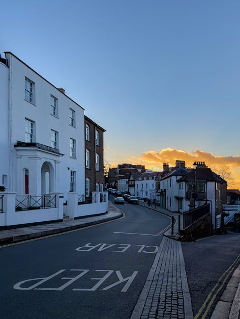 A quiet street scene in a residential area at sunset, with a narrow, uphill asphalt road marked with 'KEEP CLEAR' in large white letters, flanked by a mix of modern and traditional terraced houses. On the left, a prominent white building features three floors, rectangular windows, and a small balcony with black metal railings. To the right, older terraced houses have white and dark brick facades, tiled roofs, and chimneys, with some structures showing bay windows and brickwork details. The pavement consists of light-colored brick tiles and a textured tactile strip along the pedestrian pathway. Street parking is visible further along the road, with a few cars parked neatly. The scene is illuminated by soft natural evening lighting, with a clear sky transitioning from blue to orange and golden clouds near the horizon, suggesting a calm atmosphere. The image visually relates to private waste handling or alternative rubbish removal options often used in local property management, hinting at the importance of efficient clearance in residential neighbourhoods, as managed by companies like House Clearance Harrow.