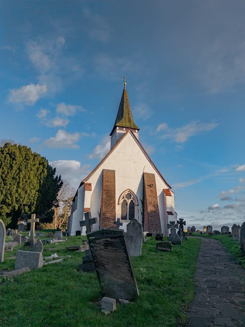 A historic white church with a steeply pitched roof and a tall, pointed steeple topped with a weather vane, set against a bright blue sky with scattered clouds. The building features narrow, arched windows and is surrounded by a graveyard with numerous old, weathered headstones of varying shapes and sizes, some leaning or tilted. The graveyard is bordered by a grassy area with a gravel path running alongside it, leading toward the church. On the left side, there are large, lush trees providing partial shade. The overall scene appears calm and well-maintained, with no visible signs of recent disturbance. The image reflects an environment where private or alternative waste handling, such as clearing debris around historical sites, may be relevant, and it captures the setting associated with waste removal at traditional or historic landmarks, aligned with professional rubbish clearance services by companies like House Clearance Harrow.