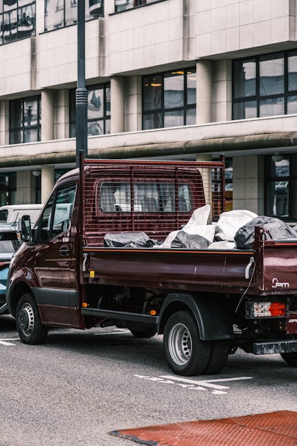 A small, dark red flatbed utility vehicle parked on an urban street in front of a modern building with large glass windows and a tiled facade. The truck's open cargo area is filled with various types of rubbish, including black garbage bags and miscellaneous debris. The vehicle features a protective metal grid at the front of the cargo bed, and the rubbish appears loosely arranged, with some bags slightly leaning or overlapping each other. Surrounding the vehicle are other parked cars and street elements like a lamppost, indicating a typical metropolitan environment. The scene suggests the vehicle is engaged in waste collection or rubbish removal, possibly for private, independent collection services, aligning with alternative waste handling options outside local authority disposal. The lighting is natural, with a diffuse daylight creating soft shadows, emphasizing the textures of the rubbish bags and the vehicle's surface, reinforcing the visual context of rubbish clearance operations.