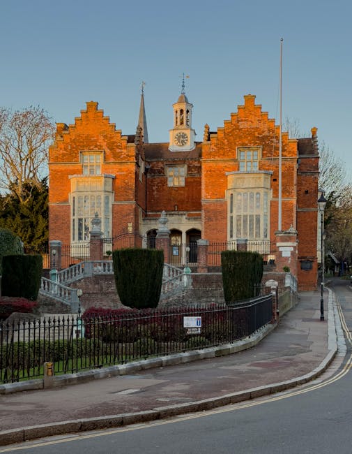 A historic brick building with ornate architectural features, including large bay windows with decorative stone surrounds, a steep gabled roof with battlements, and a prominent central clock tower with a weather vane on top. The building is situated on a slight elevation, accessed by a wide staircase with stone balustrades, surrounded by neatly trimmed bushes and a black wrought iron fence. The scene is set on a quiet street with a paved sidewalk and a lamp post on the right side. The lighting indicates late afternoon or early evening, casting warm tones on the brickwork. The setting and condition of the building suggest a property that may require occasional site clearance or specialist waste disposal, areas where companies like House Clearance Harrow may provide independent rubbish collection or on-site clearance services. The surrounding environment features a clear sky and mature trees in the background, indicating a well-maintained urban area with historical architecture.