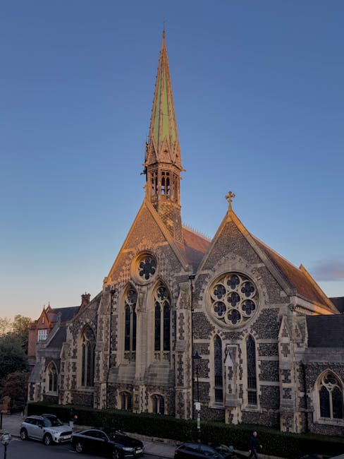 A large historic church building constructed from grey stone with intricate Gothic architectural details, including pointed arch windows with stained glass and decorative tracery, set against a clear blue sky during late afternoon or early evening. The church features a tall, slender spire with a greenish patina, topped by a small cross, rising prominently above the roofline. The façade displays smaller arched windows, buttresses, and a circular window with elaborate tracery. Surrounding the church is a paved street with parked cars and a low hedge along the boundary on the lower edge of the image, indicating an urban setting possibly near centre of a town or village. The lighting emphasizes the texture of the stonework as shadows fall across the façade, highlighting the Gothic detailing, with a few pedestrians visible at the bottom of the frame, reinforcing the building’s community and cultural significance within the local area. The scene captures the contrast between the historic architecture and the contemporary urban environment, subtly aligning with the theme of heritage preservation amidst modern waste management and local infrastructure services.